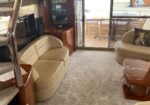 Interior of a luxury boat salon featuring a beige curved sofa, wooden glossy tables, and carpeted stairs; sliding glass doors open to a deck with wicker chairs.