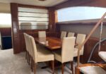 Luxurious yacht dining area with a glossy wood table and eight cream chairs, wood paneling and curved windows nearby.