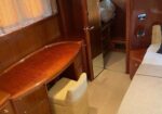 Wood-paneled boat cabin with curved varnished desk, beige chair, and mirror wardrobe near a bed.