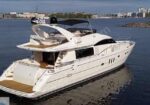 White luxury motor yacht anchored near a shoreline with calm water and a cityscape in the distance, featuring a spacious deck and flybridge.