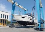 Yacht suspended in a blue dry-dock lift, being raised for maintenance near a white building and yellow machinery.