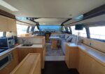 Open-plan yacht interior showing a light wood galley to the left with sink and appliances, and a seating lounge with an L-shaped sofa and central table toward the bow, large windows ahead.