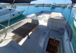 Open-air yacht deck with beige cushioned seating, a wooden table, and stairs to the cabin, docked in a marina with other boats nearby in calm water.