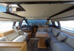 Luxurious yacht interior lounge with light gray sectional sofas around a wooden table and a flower vase near the helm.