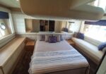 Luxurious boat cabin with a centered bed, gray bedding, and striped throw; built-in cabinetry along the left wall and cushioned seating with purple pillows on the right. Mirror wall behind bed reflects space. Porthole windows provide natural light.