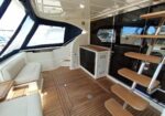 Teak-decked yacht aft lounge with built-in white seating, small galley cabinet, and a wooden ladder leading to the upper deck beside sliding glass doors