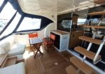 Yacht cockpit and outdoor dining area with white cushioned seating, a small table, and orange-cushioned chairs on a wood-grain deck.