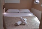 Compact cabin bedroom with a neatly made double bed, two white pillows, and rolled towels on a beige checkered bedspread, with a small porthole window nearby.