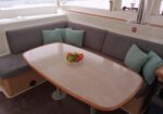 Inside a boat cabin, a U-shaped gray sofa with teal cushions surrounds a pale wooden table; a bowl of apples and oranges rests in the center.