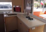 Galley kitchen on a boat with a sink, gas stove, and a shiny espresso pot on the counter, view of the outside deck through the window.