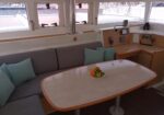 Cozy boat cabin seating area with light gray cushions and mint pillows around a wooden table with a bowl of fruit; windows show a marina outside.