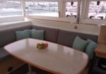 Cozy yacht cabin with a curved gray sofa, light blue cushions, and an oval wooden table holding a fruit bowl, overlooking a marina.