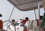 Sailboat cockpit featuring a steering wheel, winch, and neatly coiled red and white lines in a sunlit deck area and blue sky above.