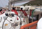 Sailboat cockpit with a large steering wheel and multiple coiled ropes on the dashboard, overlooking a marina with buildings in the background.