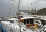 White sailing yacht docked at a marina with orange cockpit seating and blue fenders along the hull, hills in the background