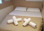 Double bed with white linens and a beige bedspread in a small cabin, four rolled white towels arranged on the duvet.