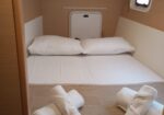 Small cabin with a neatly made bed, two pillows, and rolled towels on the floor in front; porthole window on the right and a compact door at the head of the bed.