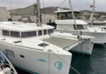 White catamaran named LARA moored at a marina, with turquoise stripe and rigging, calm water nearby.