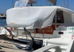 White luxury sailboat with a covered upper deck and orange seating, docked at a marina. A small inflatable dinghy sits on a cradle at the stern. The boat name LARA is visible on the side.