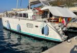 White sailing yacht moored at a marina, blue fenders along its hull and a teal stripe, open rear deck visible under a canopy.