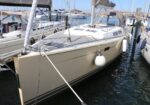 Beige sailboat moored at a busy marina with fenders on its side and other boats in the background.