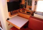 Boat cabin navigation/comms station with a small wall-mounted flat-screen monitor, multiple handheld radios, and a control panel above a light gray desk pad; red cushioned seat nearby.