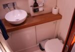 Compact bathroom with a round white vessel sink on a wooden countertop, wall faucet, shelf holding soap and a toothbrush cup, toilet to the right, and a roll of toilet paper nearby.
