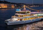 Nighttime view of a white, three‑deck passenger boat named 'Sultania' with string lights, on calm water with a lit city promenade in the background