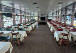 Interior of a river cruise dining hall with rows of white-tablecloth tables and colored chairs along both sides.
