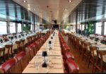 Long banquet tables set with white linens, glassware, and red-upholstered chairs in a ship's dining hall