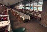 Cruise ship dining hall with rows of tables, white tablecloths, and red/green chairs by large windows overlooking the sea.