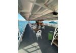 Outdoor boat deck set up for dining with white chairs and wooden tables, overlooking blue water and another boat in the distance.