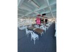 Outdoor boat dining deck with white chairs and wood tables arranged in rows, pink floral wall in background, under a blue ceiling.