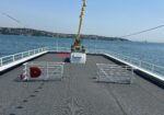 Open deck of a boat with a central cactus planter, gray non-slip surface, and white railings overlooking calm blue sea and distant shore.