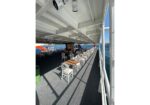 Open-air seating on a ferry deck with wooden tables and white chairs, overlooking the water.