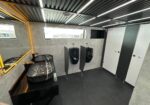 Modern men's bathroom with two wall-mounted urinals, privacy panels, and a black marble sink on a glossy black countertop beside a backlit mirror.
