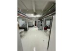 Industrial basement room with white floor and ceiling pipes; central racks, stainless tables, a red air compressor on the right, and white chairs along the walls.