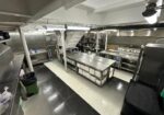 Professional stainless-steel kitchen with central prep table and stacked plates beneath, bright overhead lighting.