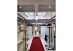 Industrial storage corridor with white machinery and racks lining both sides; a red carpet runs down the center toward a doorway at the far end.