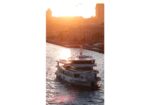 Sunset over a river with a white tour boat cruising past a city waterfront, casting a golden glow on the water.