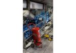 Blue industrial generator in a ship's engine room, with a red wheeled fire extinguisher cart in the foreground and yellow-handled valves nearby.