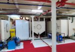 Ship engine room with large white machinery, red and blue pipes overhead, and an accessible yellow oil spill kit nearby.