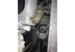 Inside a compact mechanical cabinet showing a rusted curved pipe connected by clamps, with other hoses and a diamond-plate wall on the right.