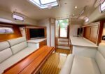 Sailboat cabin with light gray sofa, wooden dining table, and built-in TV opposite a kitchenette and cabinets.