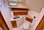 Compact marine head with cream countertop, stainless sink, wooden cabinets, and a small porthole window.