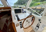 Boat cockpit with a wooden steering wheel, navigation screens, and a white control console, rocky shoreline visible through the windows.