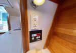 Wood-paneled interior corner with a white electrical outlet and a black control panel on a white wall, under a small ceiling light on a boat or RV galley.