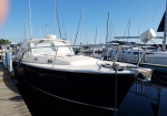 A white and black motor yacht moored at a wooden marina dock with sailboats and masts in the background.