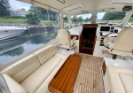Interior of a yacht cabin: cream leather sofa and wooden table beside large windows looking out to a calm waterway and another boat docked nearby; helm area with steering wheel and navigation screens to the front.