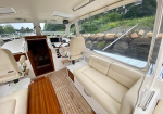 Cozy boat cockpit with beige seating, wooden table, and helm near large windows overlooking a rocky shoreline.